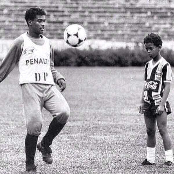 Młody Ronaldinho w czarno-białej koszulce Coca-Cola patrzy na starszego brata podbijającego piłkę
