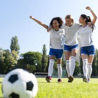 Trzy młode kobiety: Murzynka, Biała i Azjatka w strojach piłkarskich kroczą zwycięsko po murawie boiska w stronę widza i piłki na murawie