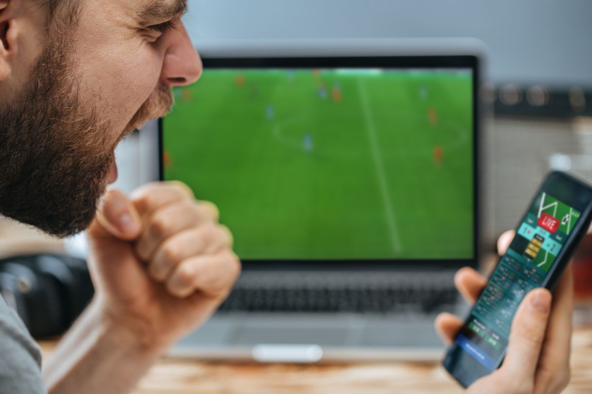 Kibic patrzy na zakłady na żywo na telefonie ze spotkaniem piłki nożnej na laptopie w tle