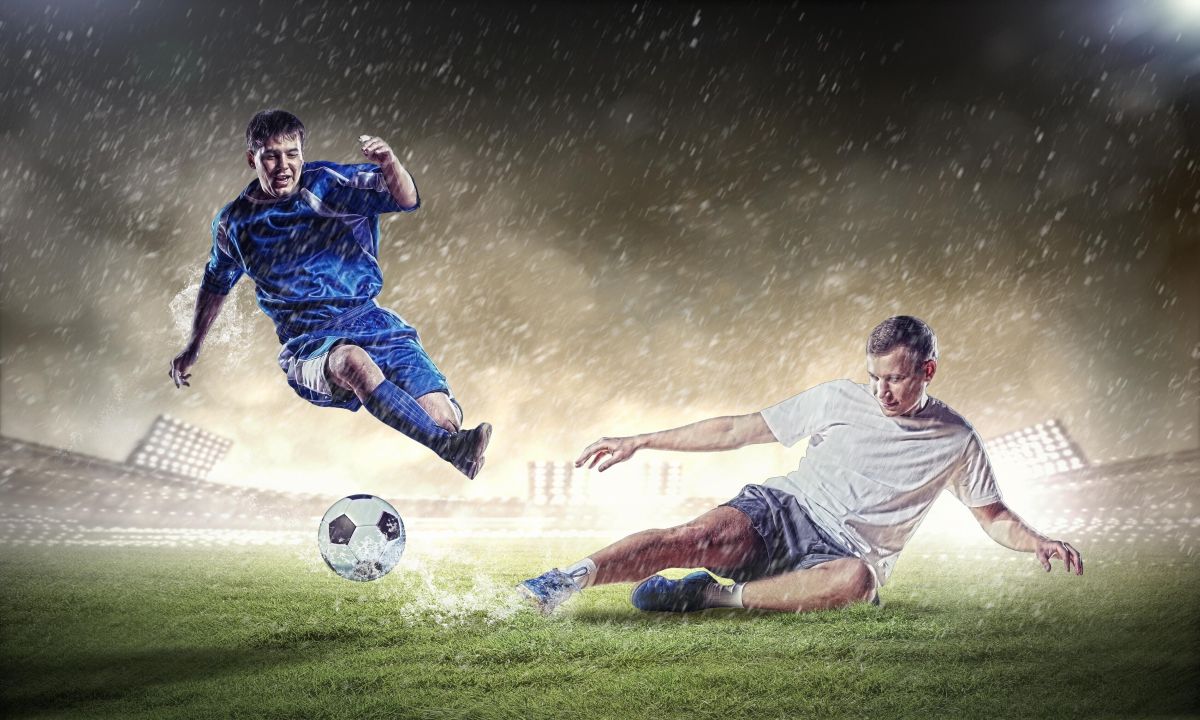Niebieski piłkarz przeskakuje nad białym piłkarzem robiącym wślizg na boisku piłkarskim na tle stadionu i zachmurzonego nieba