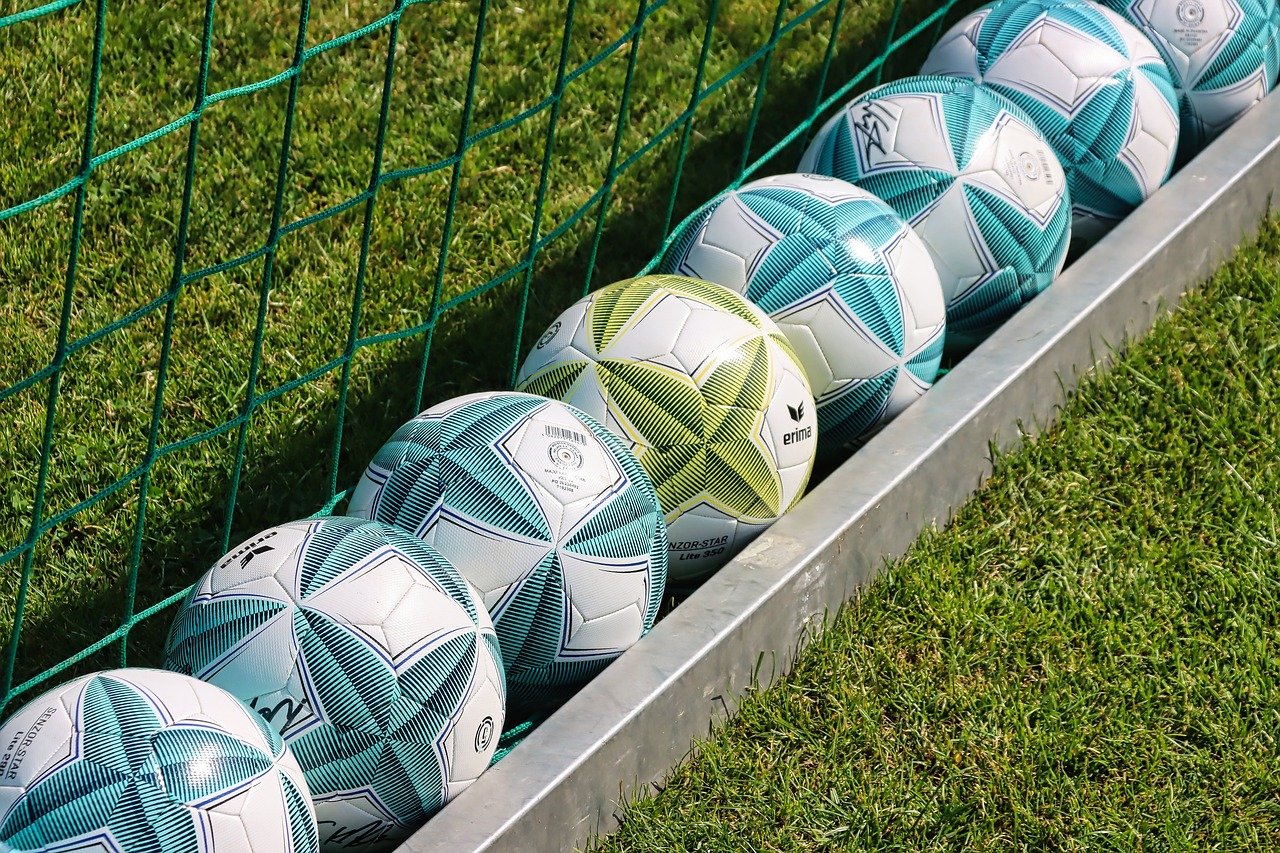 Piłki ułożone skośnie na zielonym boisku pomiędzy siatką a metalową prowadnicą